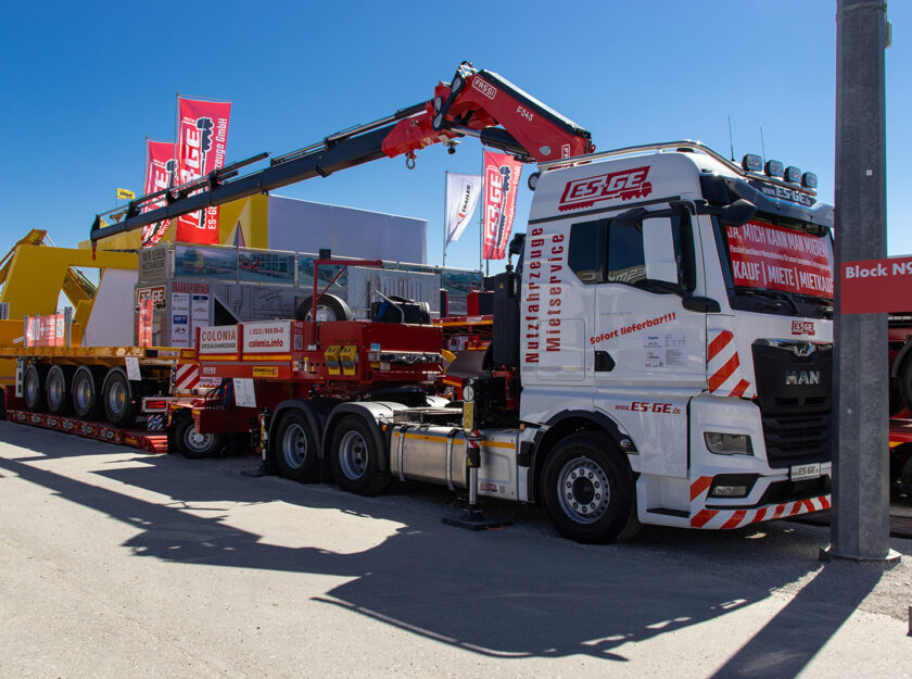 MAN Sattelzugmaschine mit Ladekran von FASSI auf dem Messestand der ES-GE auf der BAUMA 2025. Dahinter ein GigaMAX Tiefbettauflieger von Faymonville mit einem Ballastauflieger auf der Ladefläche