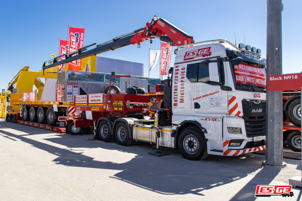 MAN Sattelzugmaschine mit Ladekran von FASSI auf dem Messestand der ES-GE auf der BAUMA 2025. Dahinter ein GigaMAX Tiefbettauflieger von Faymonville für die Firma Colonia Spezialfahrzeuge mit einem Ballastauflieger auf der Ladefläche.