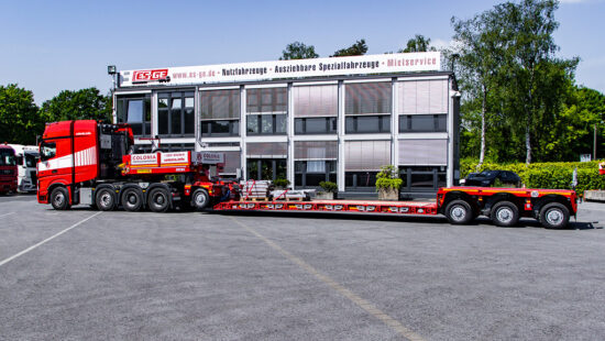 Mercedes-Benz Sattelzugmaschine von Colonia Spezialfahrzeuge mit 4 Achsen. Dahinter eine Tiefbettkombination des Typs Faymoville GigaMAX 1+3. Im Hintergrund das Gebäude der ES-GE. Ansicht des kompletten Zuges von der Fahrerseite.